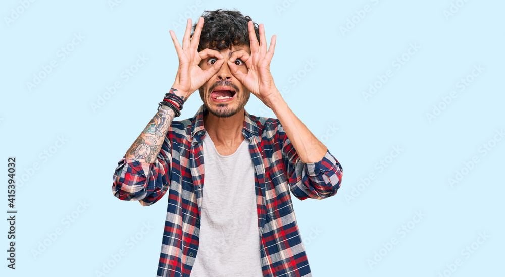Young hispanic man wearing casual clothes doing ok gesture like binoculars sticking tongue out, eyes looking through fingers. crazy expression. Young hispanic man wearing casual clothes doing ok gesture like binoculars sticking tongue out, eyes looking through fingers. crazy expression.