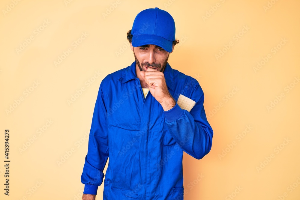 Handsome young man with curly hair and bear wearing builder jumpsuit uniform feeling unwell and coughing as symptom for cold or bronchitis. health care concept.