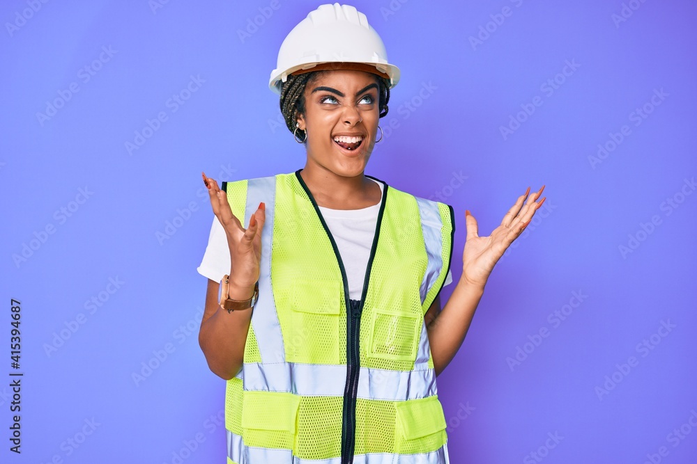 Young african american woman with braids wearing safety helmet and reflective jacket crazy and mad shouting and yelling with aggressive expression and arms raised. frustration concept. Young african american woman with braids wearing safety helmet and reflective jacket crazy and mad shouting and yelling with aggressive expression and arms raised. frustration concept.