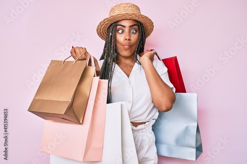 Young african american woman with braids holding shopping bags making fish face with mouth and squinting eyes, crazy and comical.