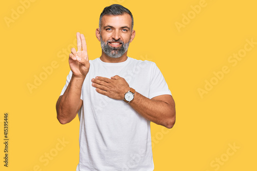 Middle age handsome man wearing casual white tshirt smiling swearing with hand on chest and fingers up, making a loyalty promise oath