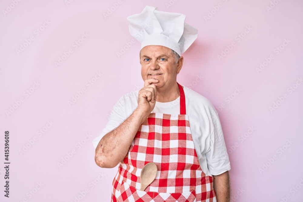 Senior grey-haired man wearing professional baker apron looking confident at the camera with smile with crossed arms and hand raised on chin. thinking positive. Senior grey-haired man wearing professional baker apron looking confident at the camera with smile with crossed arms and hand raised on chin. thinking positive.