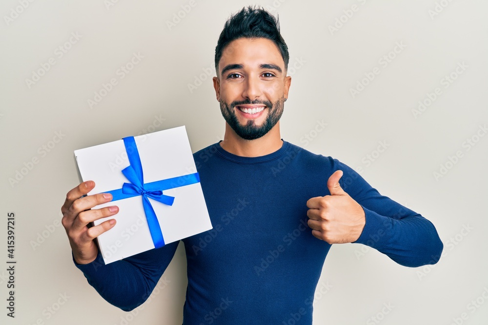 Young man with beard holding gift smiling happy and positive, thumb up doing excellent and approval sign Young man with beard holding gift smiling happy and positive, thumb up doing excellent and approval sign