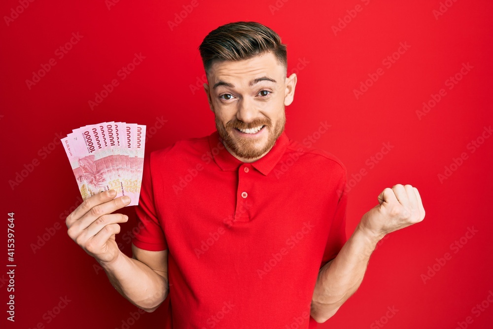 Young redhead man holding 100000 indonesian rupiah screaming proud, celebrating victory and success very excited with raised arm Young redhead man holding 100000 indonesian rupiah screaming proud, celebrating victory and success very excited with raised arm