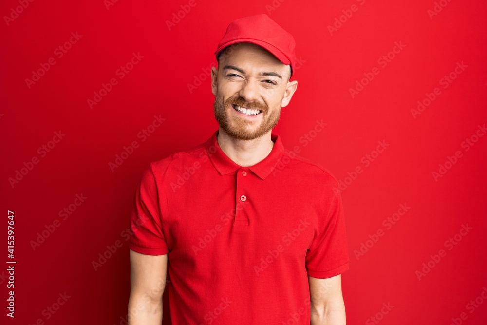 Young redhead man wearing delivery uniform and cap looking positive and happy standing and smiling with a confident smile showing teeth