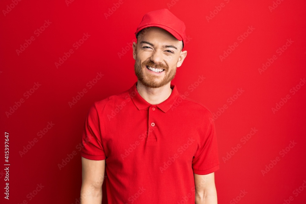 Young redhead man wearing delivery uniform and cap looking positive and happy standing and smiling with a confident smile showing teeth Young redhead man wearing delivery uniform and cap looking positive and happy standing and smiling with a confident smile showing teeth