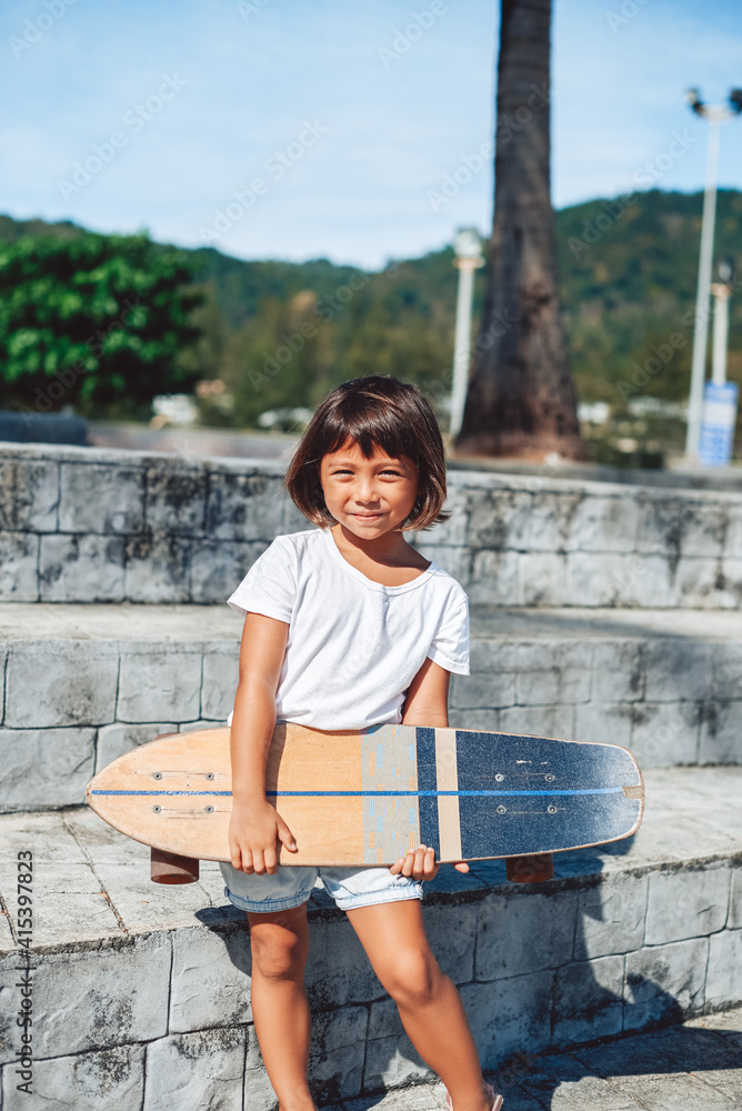 Sportive lifestyle of a little girl in park. Youth girl dressed in shorts with white shirt stays on stairs holding a skateboard outdoors.