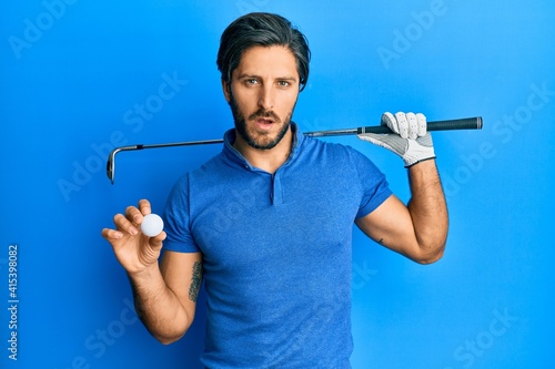 Young hispanic man holding golf ball in shock face, looking skeptical and sarcastic, surprised with open mouth