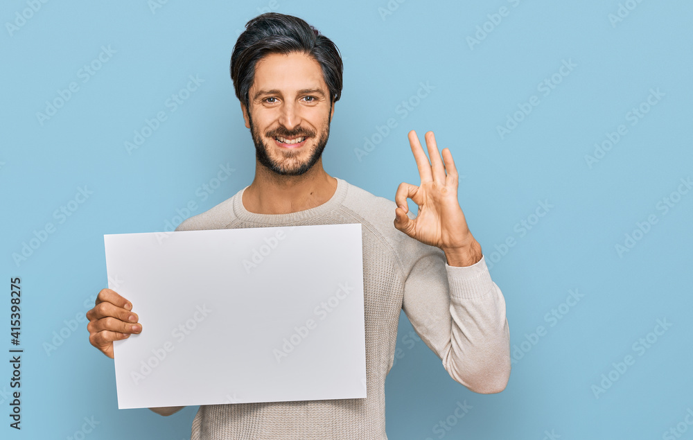 Young hispanic man holding blank empty banner doing ok sign with fingers, smiling friendly gesturing excellent symbol Young hispanic man holding blank empty banner doing ok sign with fingers, smiling friendly gesturing excellent symbol