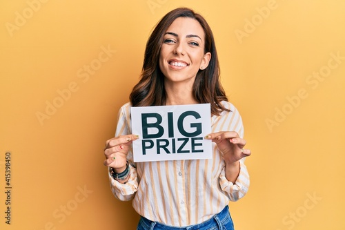 Young brunette woman holding big prize paper looking positive and happy standing and smiling with a confident smile showing teeth
