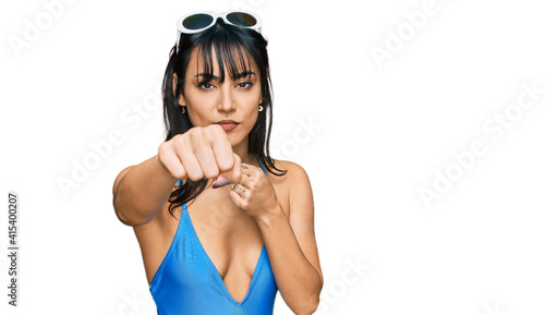 Young hispanic woman wearing swimsuit and sunglasses punching fist to fight, aggressive and angry attack, threat and violence