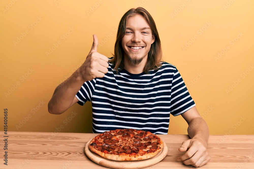 Handsome caucasian man with long hair eating tasty pepperoni pizza smiling happy and positive, thumb up doing excellent and approval sign Handsome caucasian man with long hair eating tasty pepperoni pizza smiling happy and positive, thumb up doing excellent and approval sign