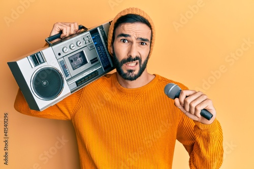 Young hispanic man holding boombox, listening to music singing with microphone clueless and confused expression. doubt concept.