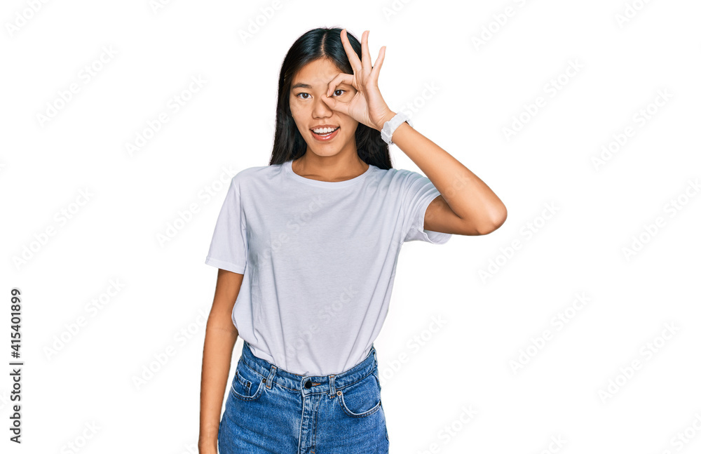Beautiful young asian woman wearing casual white t shirt doing ok gesture with hand smiling, eye looking through fingers with happy face. Beautiful young asian woman wearing casual white t shirt doing ok gesture with hand smiling, eye looking through fingers with happy face.