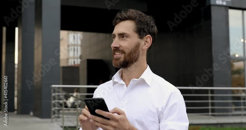 Confident bearded man browsing internet on modern smartphone while standing near office center. Happy man taking break from work on fresh air.
