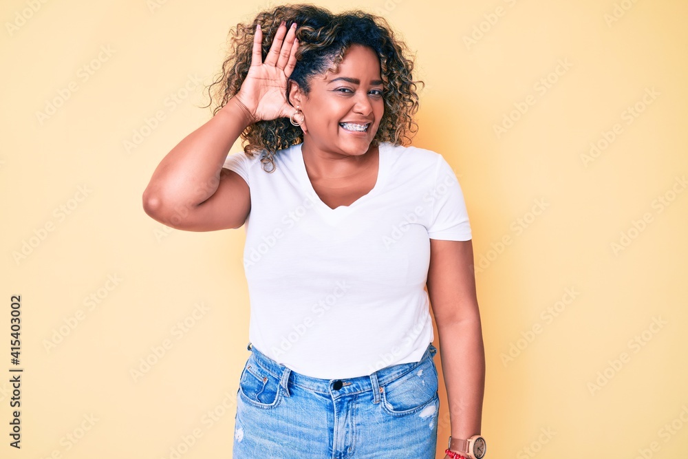 Young african american plus size woman wearing casual white tshirt smiling with hand over ear listening an hearing to rumor or gossip. deafness concept. Young african american plus size woman wearing casual white tshirt smiling with hand over ear listening an hearing to rumor or gossip. deafness concept.