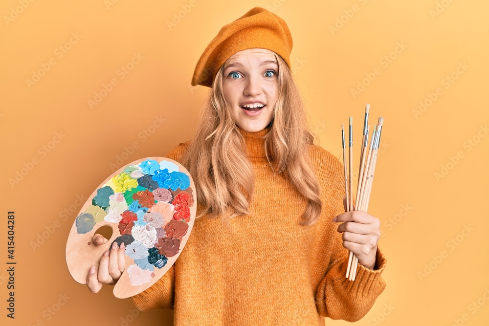Fototapeta premium Beautiful young caucasian girl holding paintbrush and painter palette celebrating crazy and amazed for success with open eyes screaming excited.