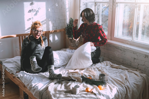 Two young girls fight pillows on the bed in a white room next to the window. Friends or sisters relationships at home