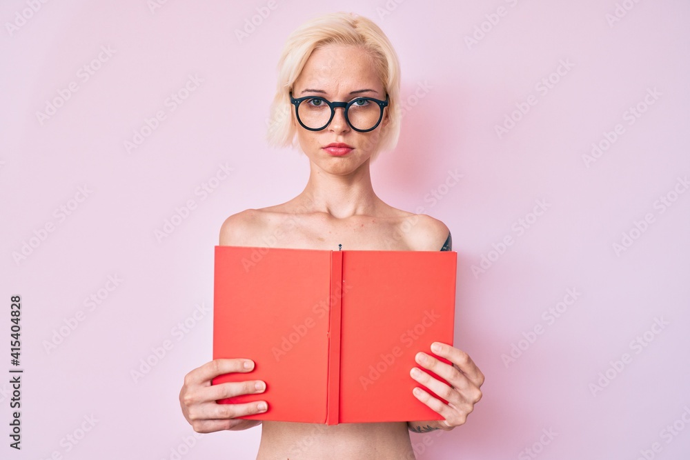 Young blonde woman with tattoo standing shirtless reading book depressed and worry for distress, crying angry and afraid. sad expression. Young blonde woman with tattoo standing shirtless reading book depressed and worry for distress, crying angry and afraid. sad expression.
