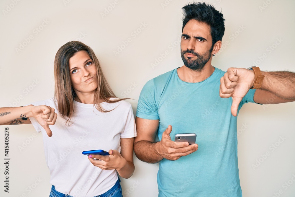 Beautiful young couple of boyfriend and girlfriend using smartphone with angry face, negative sign showing dislike with thumbs down, rejection concept Beautiful young couple of boyfriend and girlfriend using smartphone with angry face, negative sign showing dislike with thumbs down, rejection concept