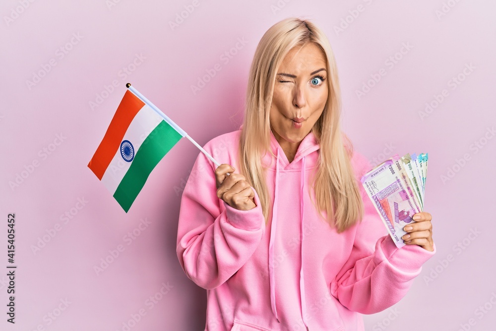 Young blonde woman holding india flag and rupee banknotes making fish face with mouth and squinting eyes, crazy and comical. Young blonde woman holding india flag and rupee banknotes making fish face with mouth and squinting eyes, crazy and comical.