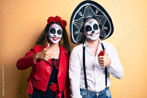 Couple wearing day of the dead costume over yellow doing happy thumbs up gesture with hand. approving expression looking at the camera showing success.
