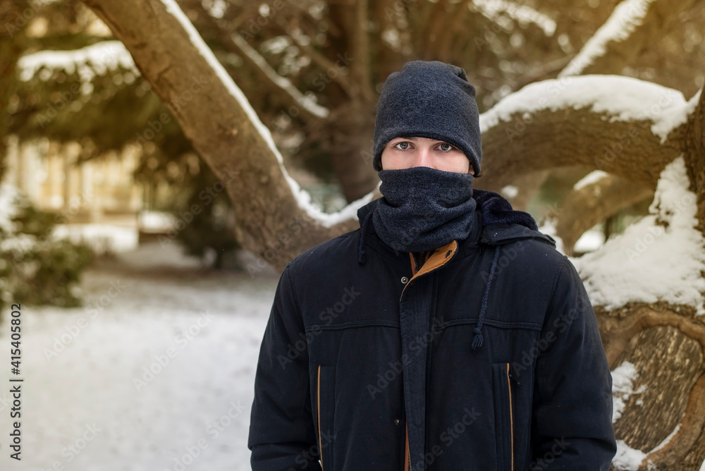 The teenager in warm clothes and in a warm hat and scarf covered half of his face because of severe frosts and snow around, the guy is sad because he loves the heat, summer