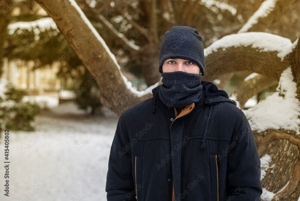 The teenager in warm clothes and in a warm hat and scarf covered half of his face because of severe frosts and snow around, the guy is sad because he loves the heat, summer The teenager in warm clothes and in a warm hat and scarf covered half of his face because of severe frosts and snow around, the guy is sad because he loves the heat, summer