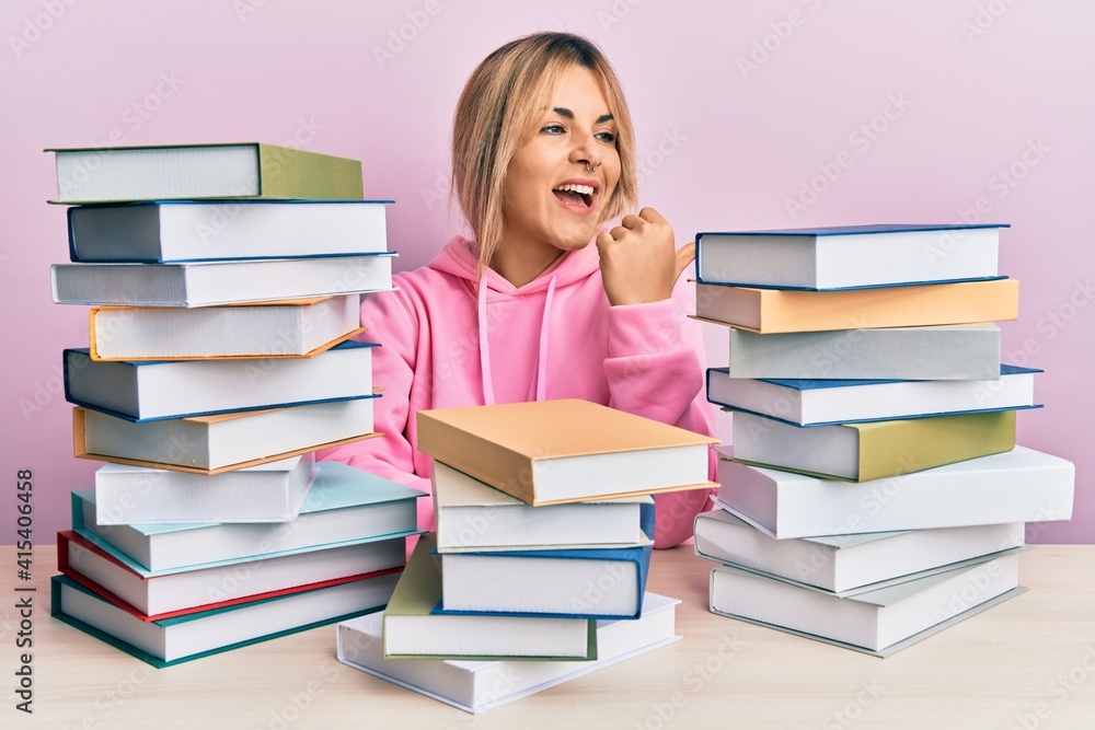 Young caucasian woman sitting on the table with books smiling with happy face looking and pointing to the side with thumb up. Young caucasian woman sitting on the table with books smiling with happy face looking and pointing to the side with thumb up.