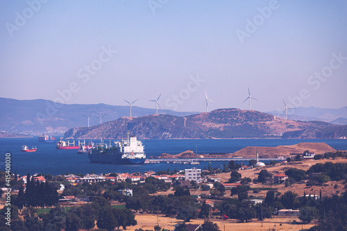 Ships are parked in a town called Aliaga in Izmir, Turkey. rotating blades of windmill propellers on blue sky background. Wind power generation. Pure green energy. Industrial city view from distance.