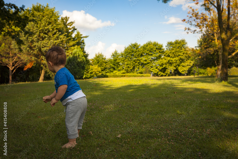 1 year old little adorable baby boy is trying to stand still in a park with bare foods on a beautiful day with some clouds. Childhood and first steps concept.