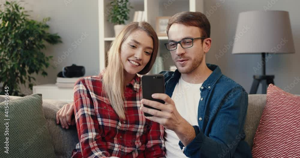 Happy husband shows photo on phone to his wife sitting at home on couch. Young married couple spending free time together. Happy husband shows photo on phone to his wife sitting at home on couch. Young married couple spending free time together.
