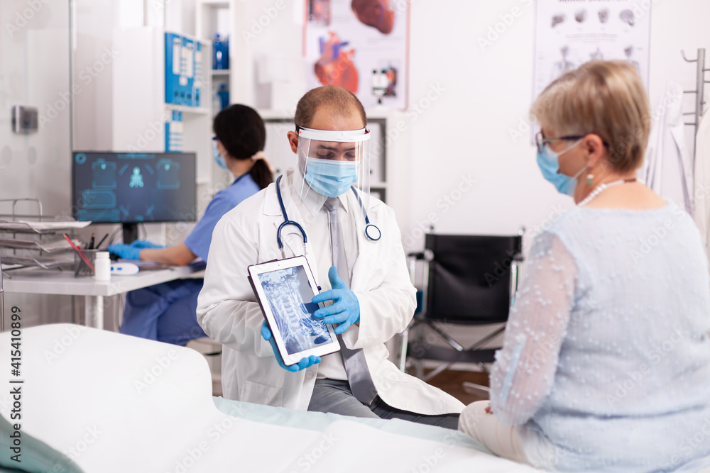 Medic explaining treatment to senior woman pointing at x-ray on tablet pc wearing face mask. Medical physician specialist during coronavirus outbreak discussing with patient.