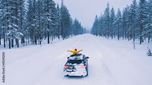 Lapland / Finland - 01.12.2020: Bird's eye view, female tourist into hatch of car raised arms enjoying winter trip in north country while driving on snowy road surrounded by coniferous forest
