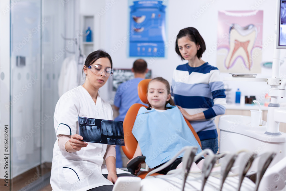 Parent and kid in dentistiry office looking at dental radiography holded by dentist. Stomatologist explaining teeth diagnosis to mother of child in health clinic holding x-ray. Parent and kid in dentistiry office looking at dental radiography holded by dentist. Stomatologist explaining teeth diagnosis to mother of child in health clinic holding x-ray.