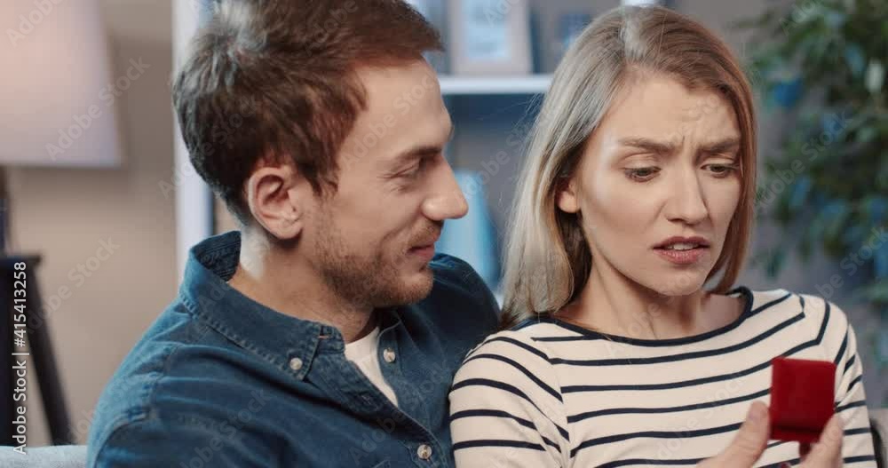 Young couple sitting at home on couch. Man gives woman box with wedding ring, dissatisfied woman refuses him. Young couple sitting at home on couch. Man gives woman box with wedding ring, dissatisfied woman refuses him.