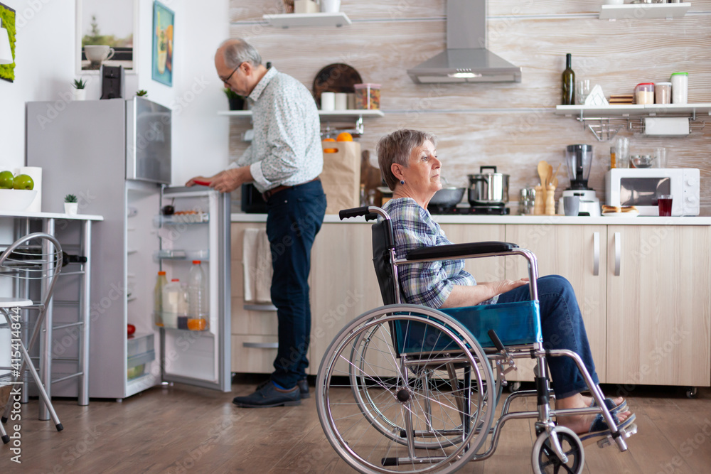 Handicapped senior woman in wheelchair sitting in kitchen looking throug window while husband unpacking grocery bag. Old disabled lady after injury and rehab, depressed invalid full of sorrow