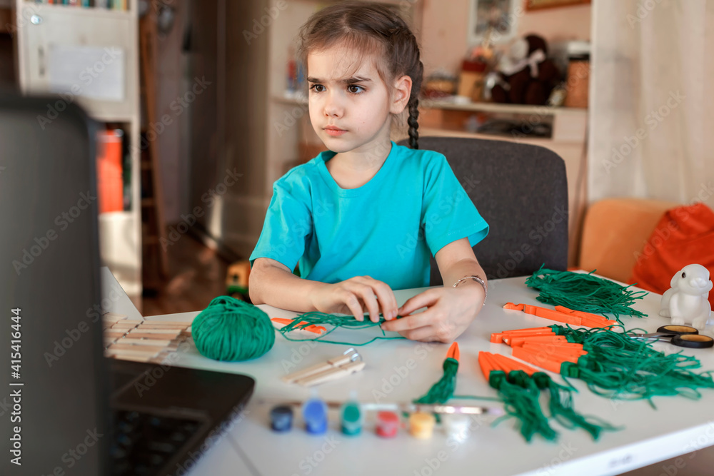 Fototapeta premium Elementary school girl doing carrot with wooden clothes pins for Easter celebration looking video on laptop, online workshop, kid at-home activity, creativity and distant education, holiday decoration