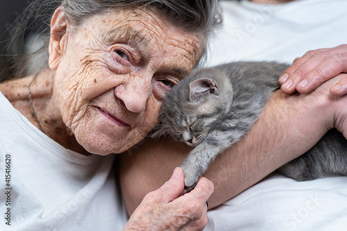Senior woman tenderness, kisses cute gray Scottish Straight kitten on couch at nursing home with volunteer. Kitty therapy. Grandmother and adult grandson stroking, spending time together with pet