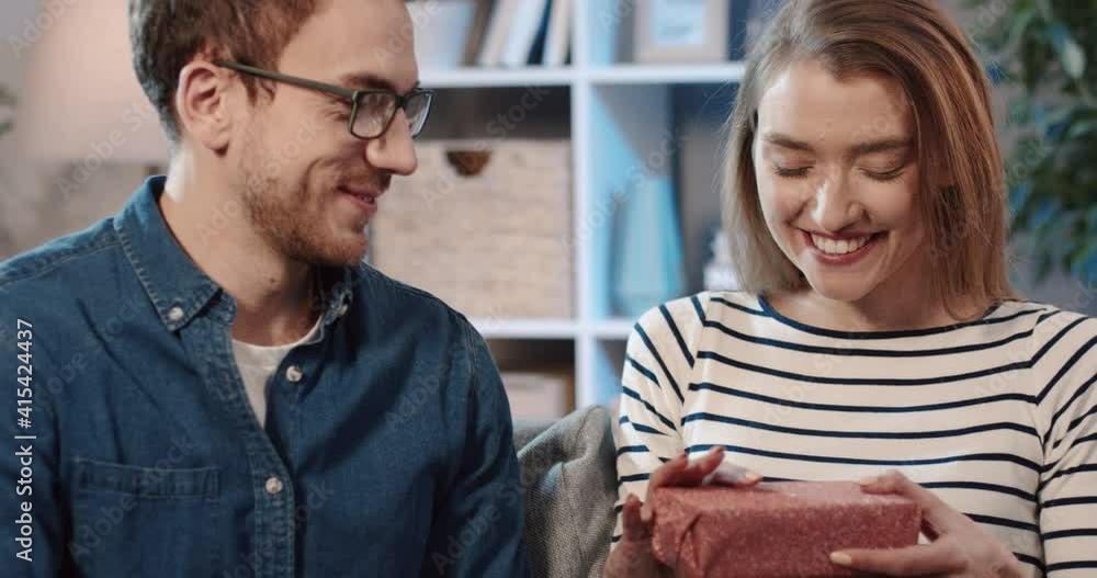 Handsome man makes gift to his wife by giving her packed box. Happy woman surprised and shakes gift in her hands. Handsome man makes gift to his wife by giving her packed box. Happy woman surprised and shakes gift in her hands.