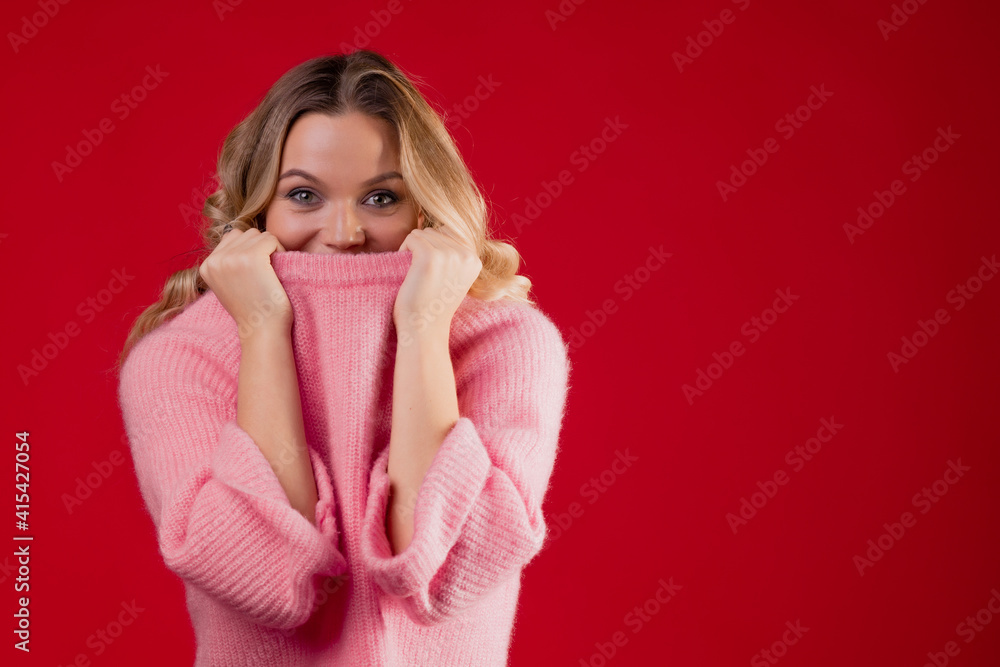 A young sweet and smiling blonde woman in a pink knitted sweater on a red background, wrapped in a collar A young sweet and smiling blonde woman in a pink knitted sweater on a red background, wrapped in a collar