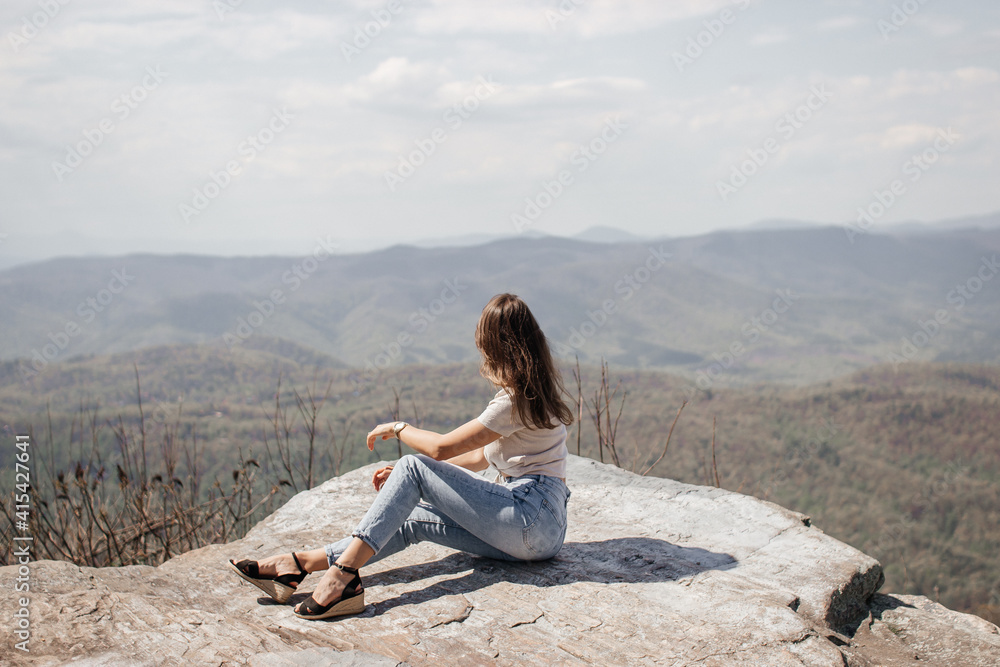 A beautiful girl with brown curly hair, in a linen short blouse with a knot and jeans, sits on a bald stone and looks into the mountains on a sunny spring day.