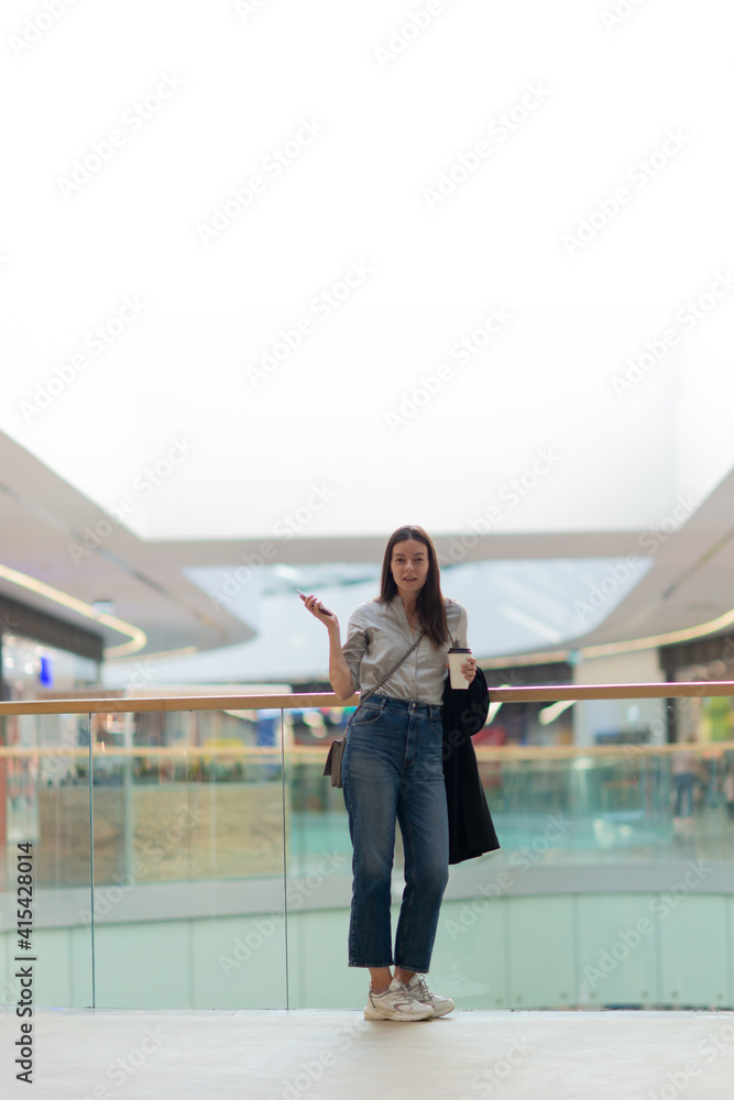 Young woman in jeans and casual clothes in a shopping center. A brunette woman with a phone in the atrium of the Mall, uses the phone for tracking purchases