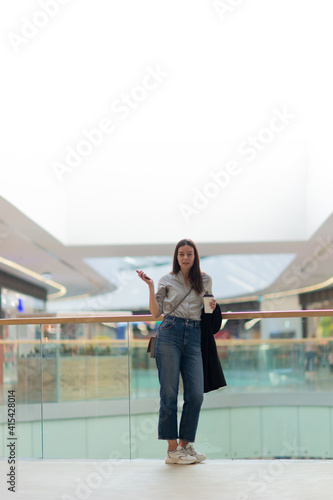 Young woman in jeans and casual clothes in a shopping center. A brunette woman with a phone in the atrium of the Mall, uses the phone for tracking purchases