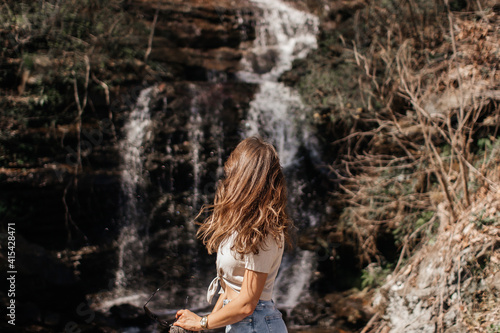 A young girl with brown curly hair in blue jeans and a linen sweater looks at a waterfall on a spring sunny day