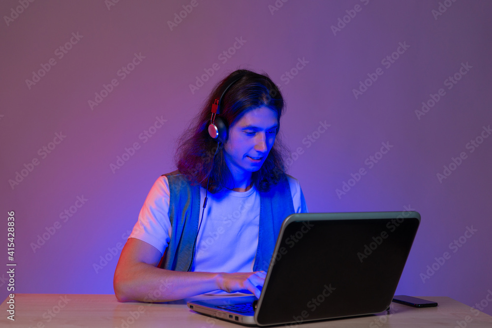 Online programming training, computer programmer or student. A young man with long hair and headphones. Use a laptop, neon tinting Online programming training, computer programmer or student. A young man with long hair and headphones. Use a laptop, neon tinting