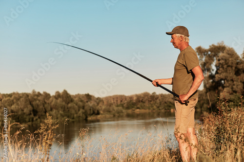 Foto Side view of fisher standing on bank of lake or river and looking at his fishing rod in hands, fishing on sunset, at beautiful nature, wearing green t shirt and trousers