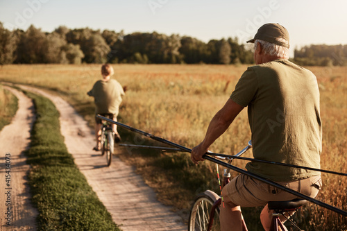 Grandfather and his grandson go fishing on bicycles, back view of family in meadow on bikes with fishing rods, senior man and young guy wearing casual closing, beautiful field and trees.