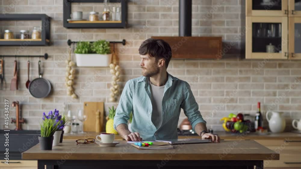 Lockdown medium shot of young man finishing remote work, closing laptop computer and looking away thoughtfully standing at table in domestic kitchen. Entrepreneur thinking of problem solution at home Lockdown medium shot of young man finishing remote work, closing laptop computer and looking away thoughtfully standing at table in domestic kitchen. Entrepreneur thinking of problem solution at home