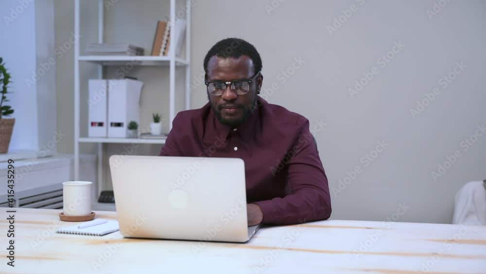 Businessman using laptop and drinking coffee while sitting at table in light office spbas. Young American man is looking at computer screen and enjoying drink at desk in modern interior. One Businessman using laptop and drinking coffee while sitting at table in light office spbas. Young American man is looking at computer screen and enjoying drink at desk in modern interior. One